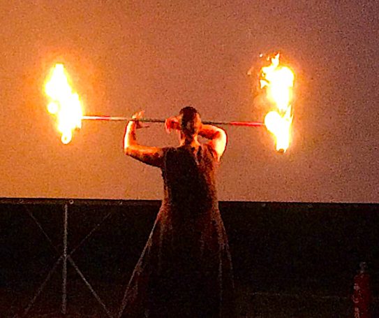 Die Glutrabe macht Kontakt-Jonglage mit einem langen Fackelstab, an dessen Enden je 3 Fackeln stehen.